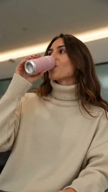 A frame from an Adapt video: a woman in a cream sweater drinks from a pink can.